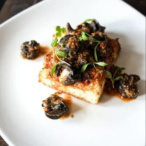 Plated gourmet dish: fried base, rich sauce, olives, microgreens.