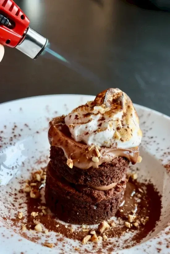 Chocolate dessert being torched with whipped cream, nuts, and chocolate sauce.