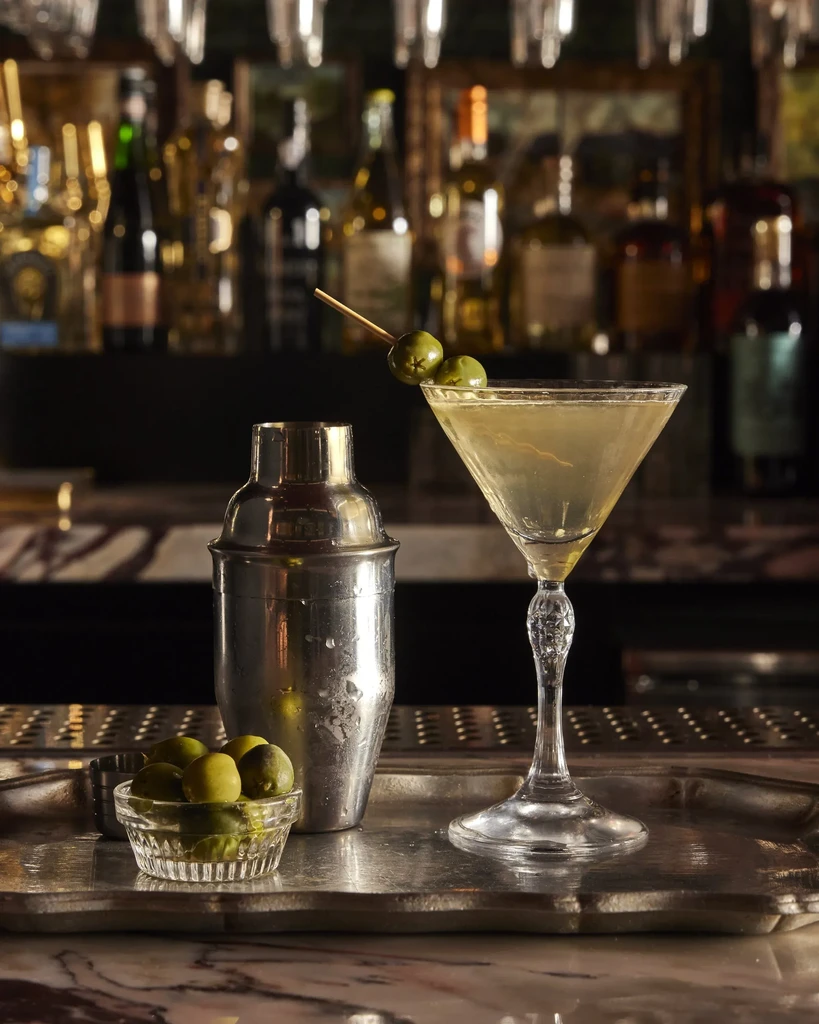 Martini, cocktail shaker, and olives on an ornate silver tray.