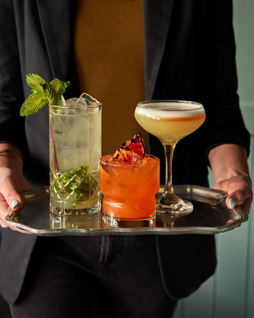 A person holding a tray of three colorful, garnished cocktails.
