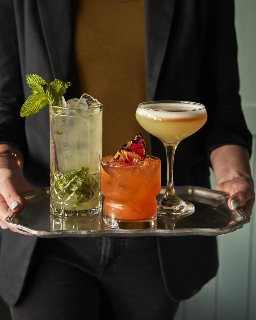 Person holding silver tray with three distinct, garnished cocktails.
