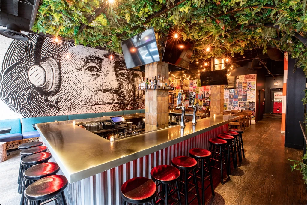 Empty eclectic bar with Benjamin Franklin mural, ceiling greenery, and red bar.