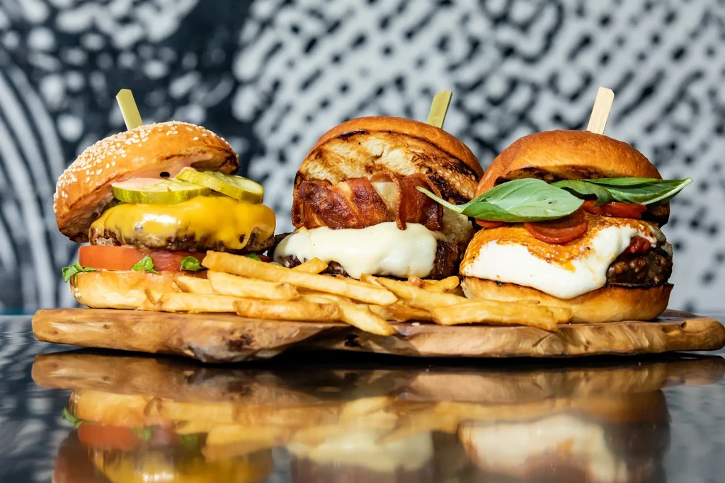 Three burgers, fries on wooden board, reflecting on dark surface.