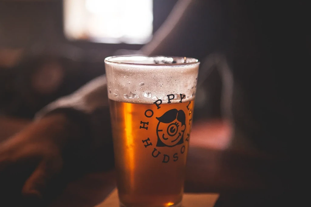 Pint of golden amber beer with 'Hoppy Hudson' logo in a dim pub.
