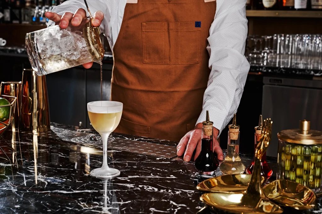 Bartender pours amber liquid from mixing glass into stemmed cocktail glass.