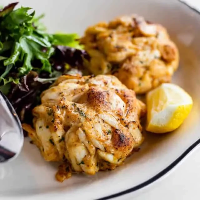 Two golden-brown crab cakes, lemon, and mixed greens on a plate.