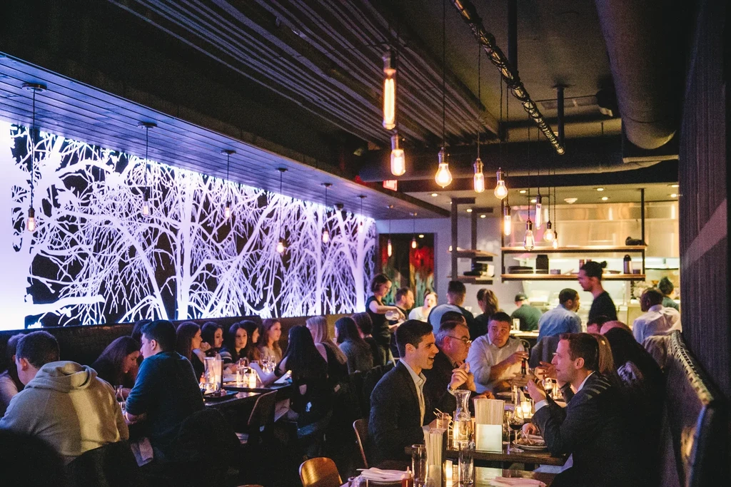 Dimly lit modern restaurant with patrons, decorative tree wall, and open kitchen.