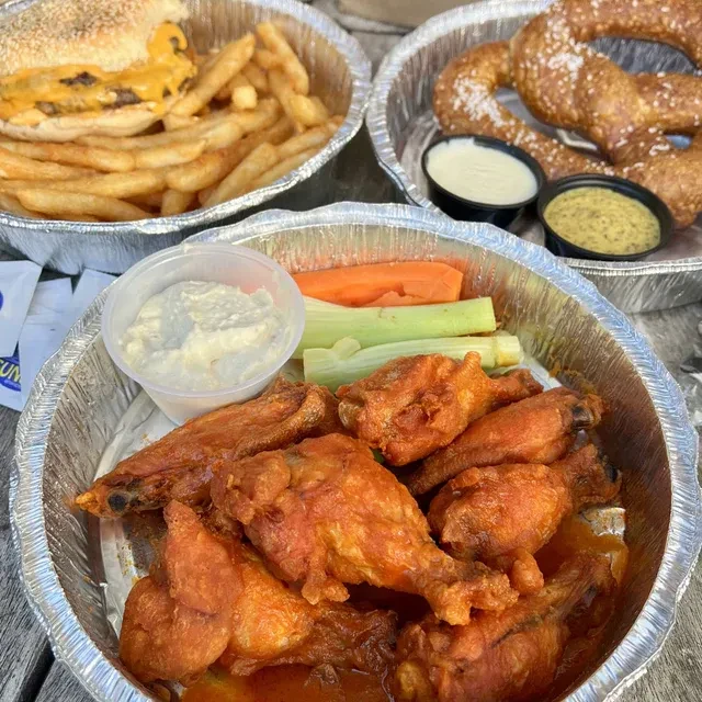 Close-up of takeout buffalo wings, cheeseburger, and pretzels in foil containers.