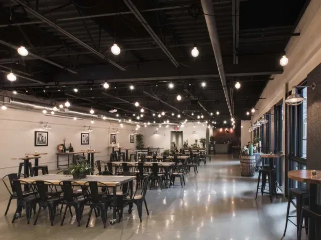 Empty industrial-chic venue interior with communal tables and bare-bulb lights.