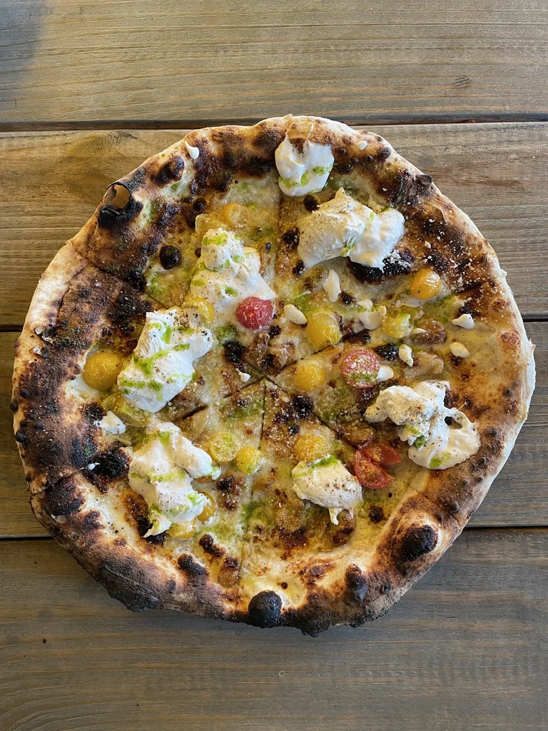 Top-down view of wood-fired pizza with burrata, tomatoes, and pesto.
