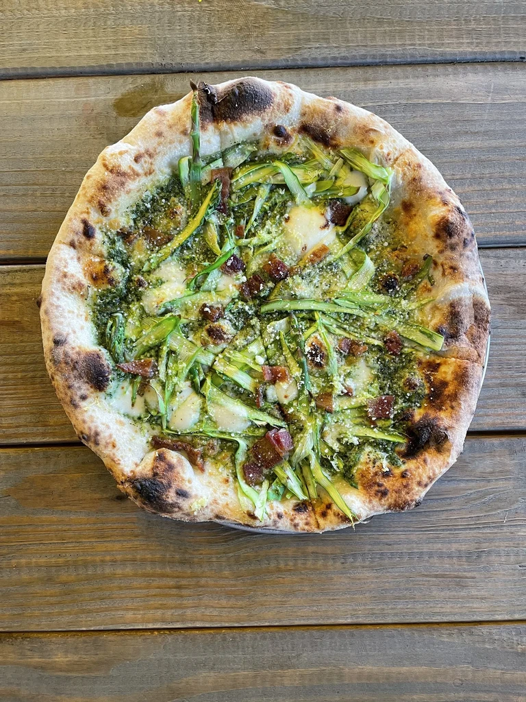 Wood-fired pizza with pesto, asparagus, bacon, and cheese on a rustic table.