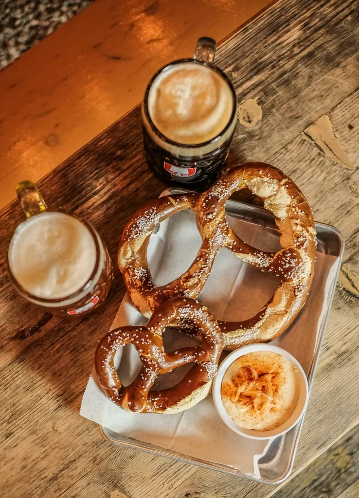 Two salted pretzels, creamy dip, and two frothy beers on a wooden table.