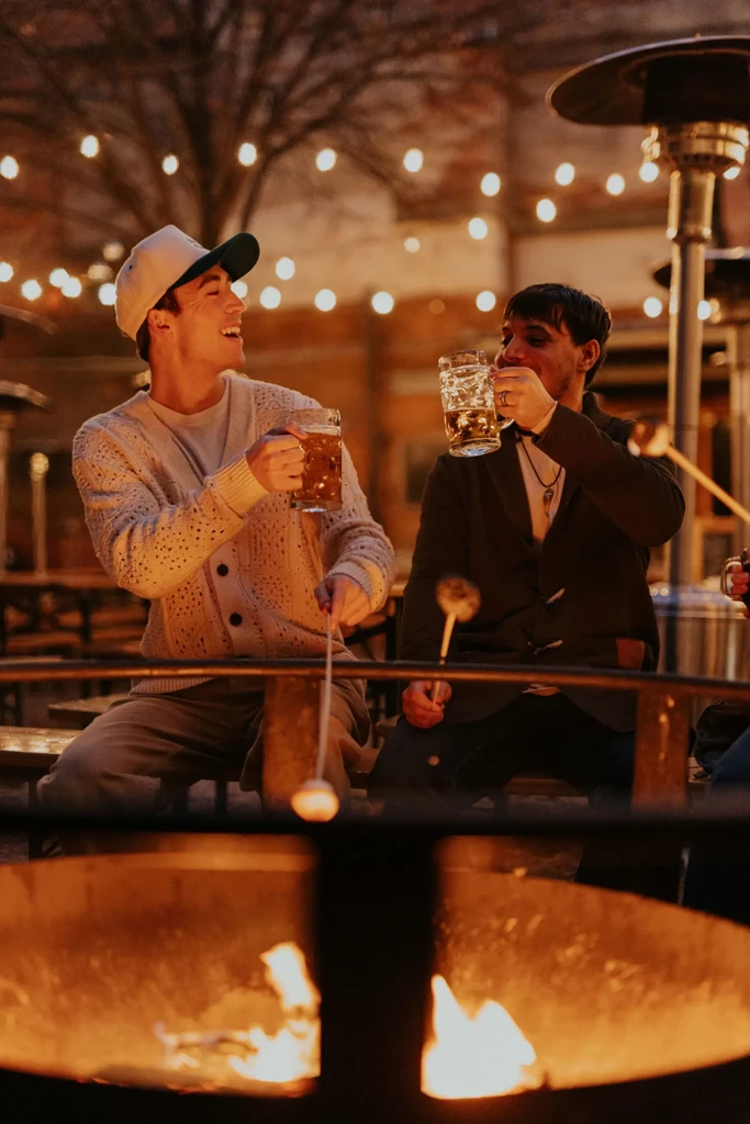Two men hold beer and roast food over an outdoor fire pit.