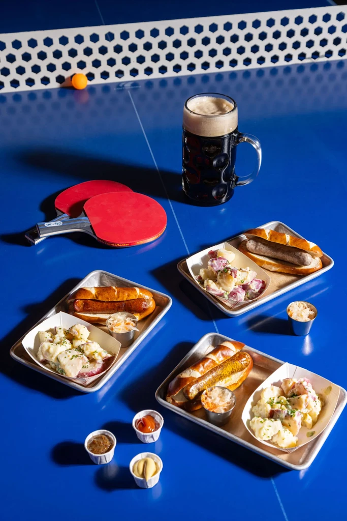 Three pretzel-bun sausages with sides and beer on a blue ping-pong table.