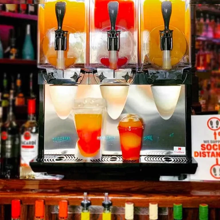 Three-tank frozen drink machine dispenses layered orange and red drinks at a bar.