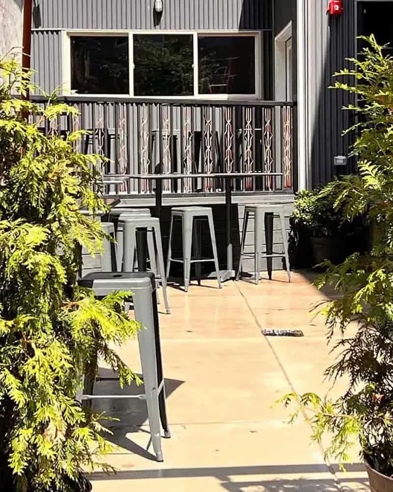 Sunny outdoor patio with high-top seating, green plants, and geometric railing.