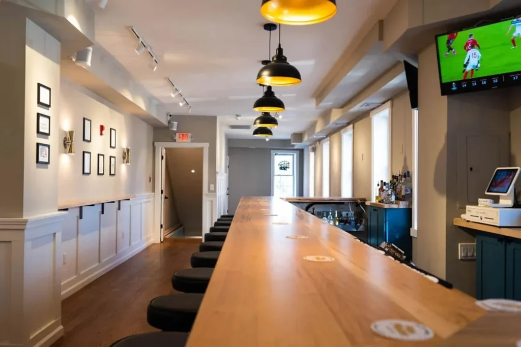 Modern bar interior with long wooden counter, stools, and a sports game.