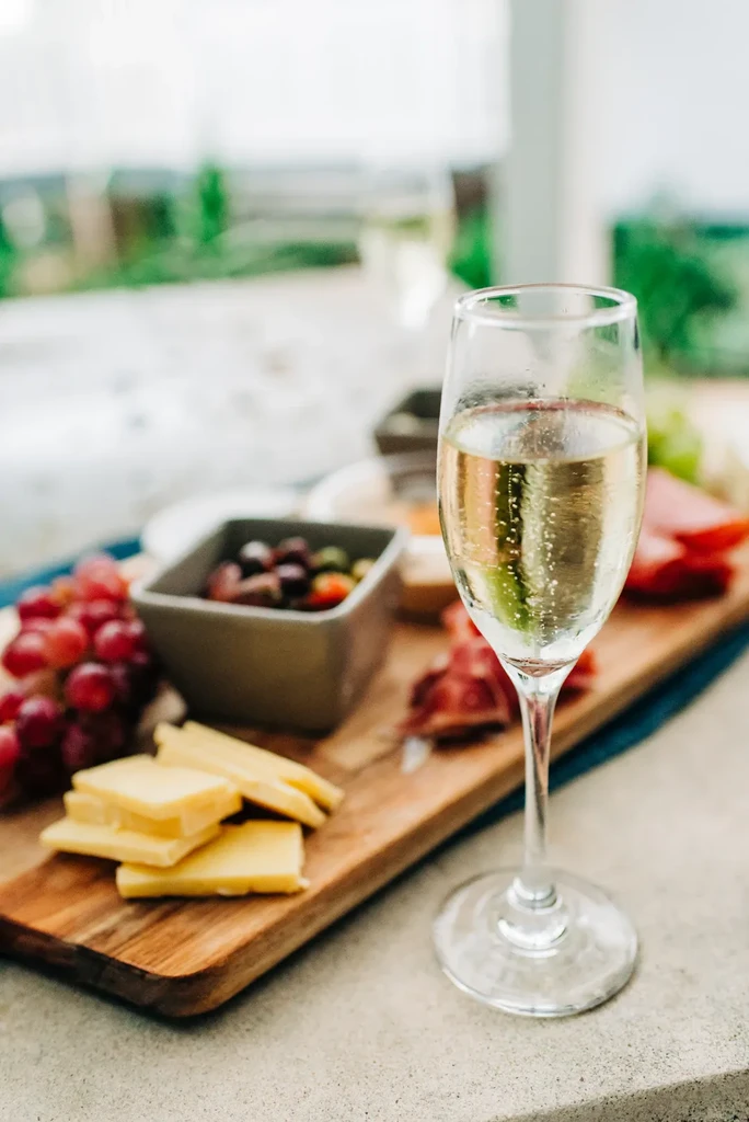 Close-up: Sparkling wine glass, wooden board with cheese, grapes, olives.