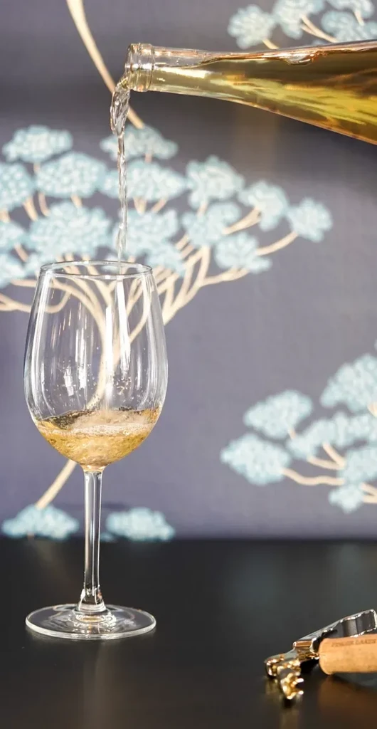 White wine being poured into a glass; patterned wall and corkscrew.