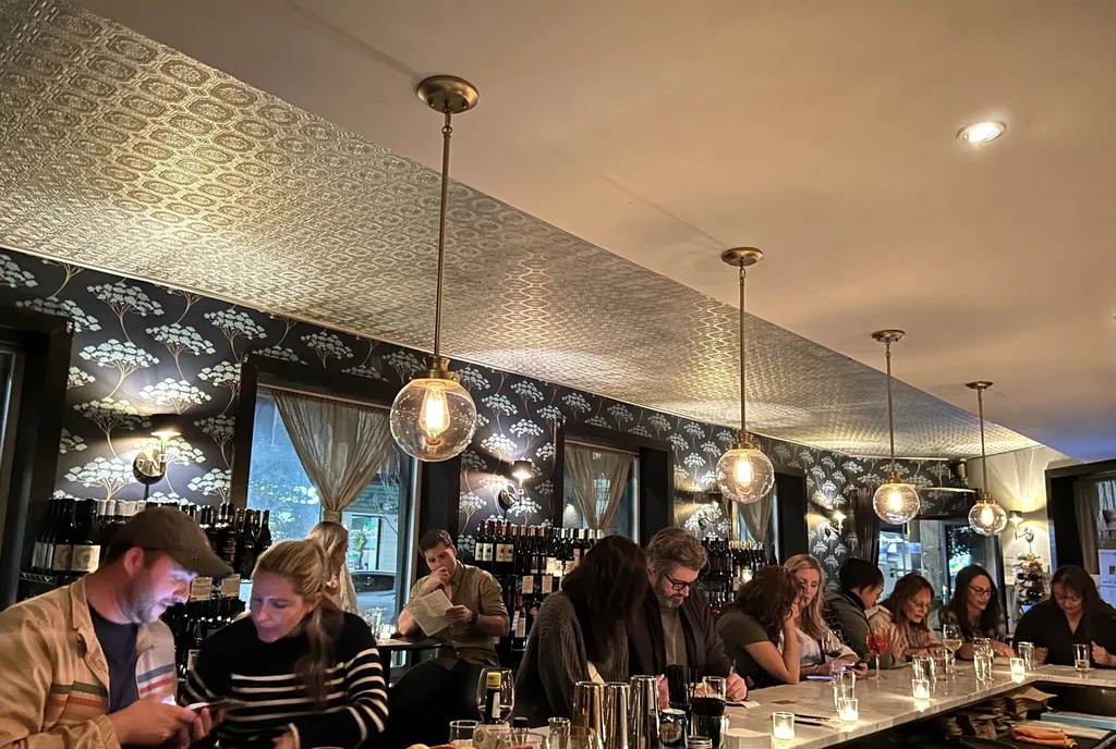 Warm, bustling bar interior with patrons, patterned decor, and wine bottles.
