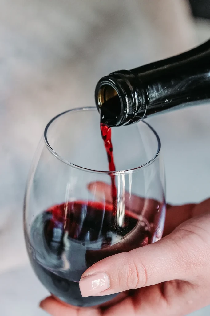 Dark red wine pouring from black bottle into a clear stemless glass.