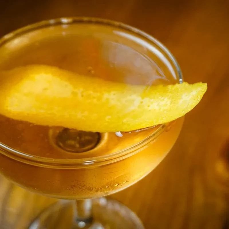 Overhead, amber cocktail in stemmed glass with yellow citrus peel garnish.