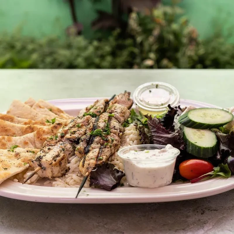 Plate of grilled chicken skewers, rice, pita, salad, and sauces.