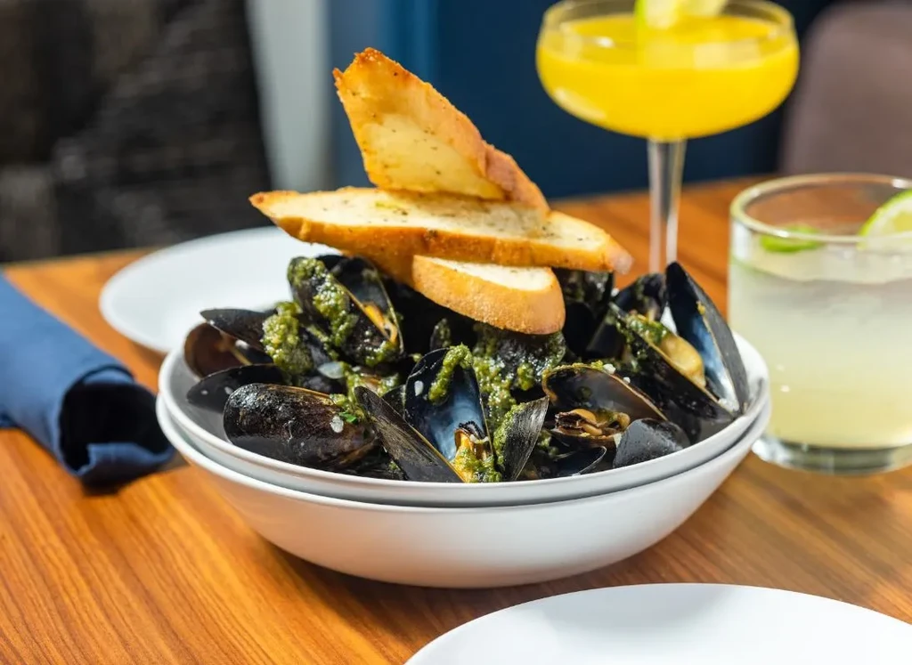 Close-up of mussels in green sauce with toast, drinks in background.