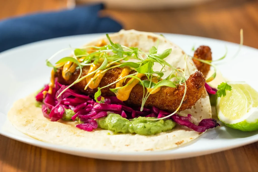 Fried fish/shrimp taco with avocado, purple cabbage, sauce, lime on white plate.