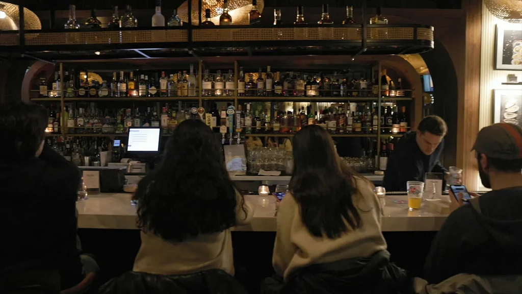Dimly lit bar interior showing customers, bartender, and many bottles.