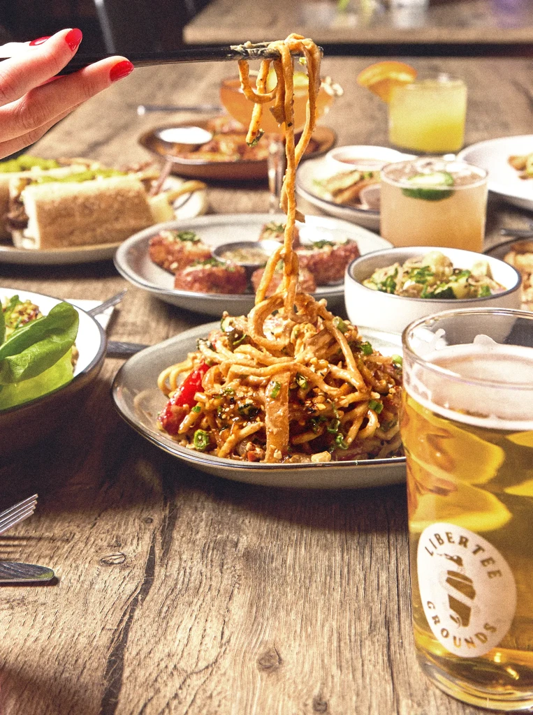 Hand lifting noodles from a table of diverse food, cocktails, and beer.