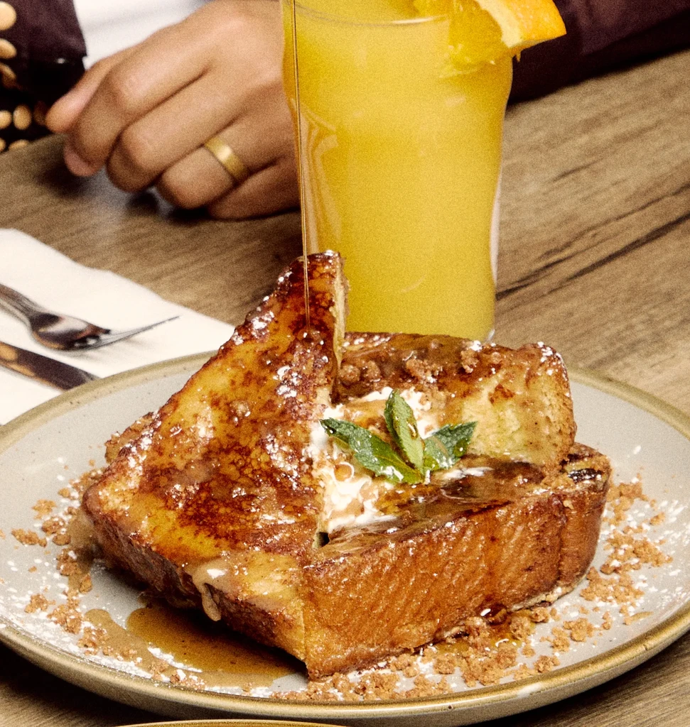Golden French toast with pouring syrup, whipped cream, mint, and orange juice.