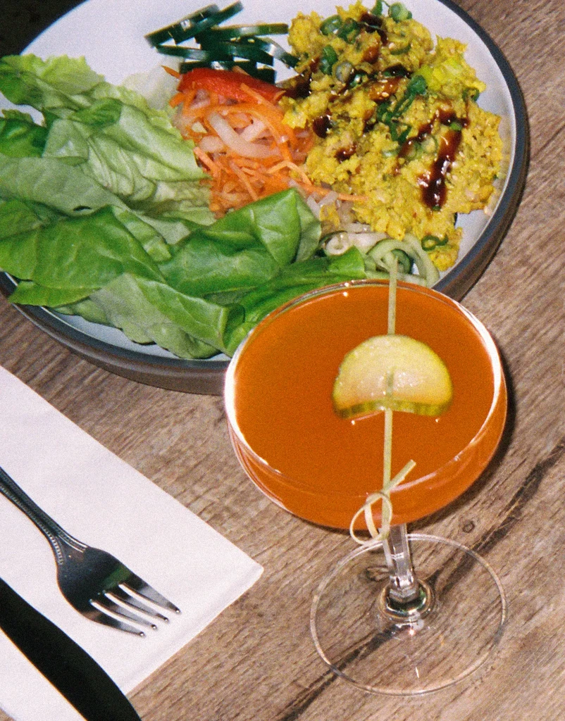 Vibrant curried bowl meal and cocktail on a light brown wooden table.