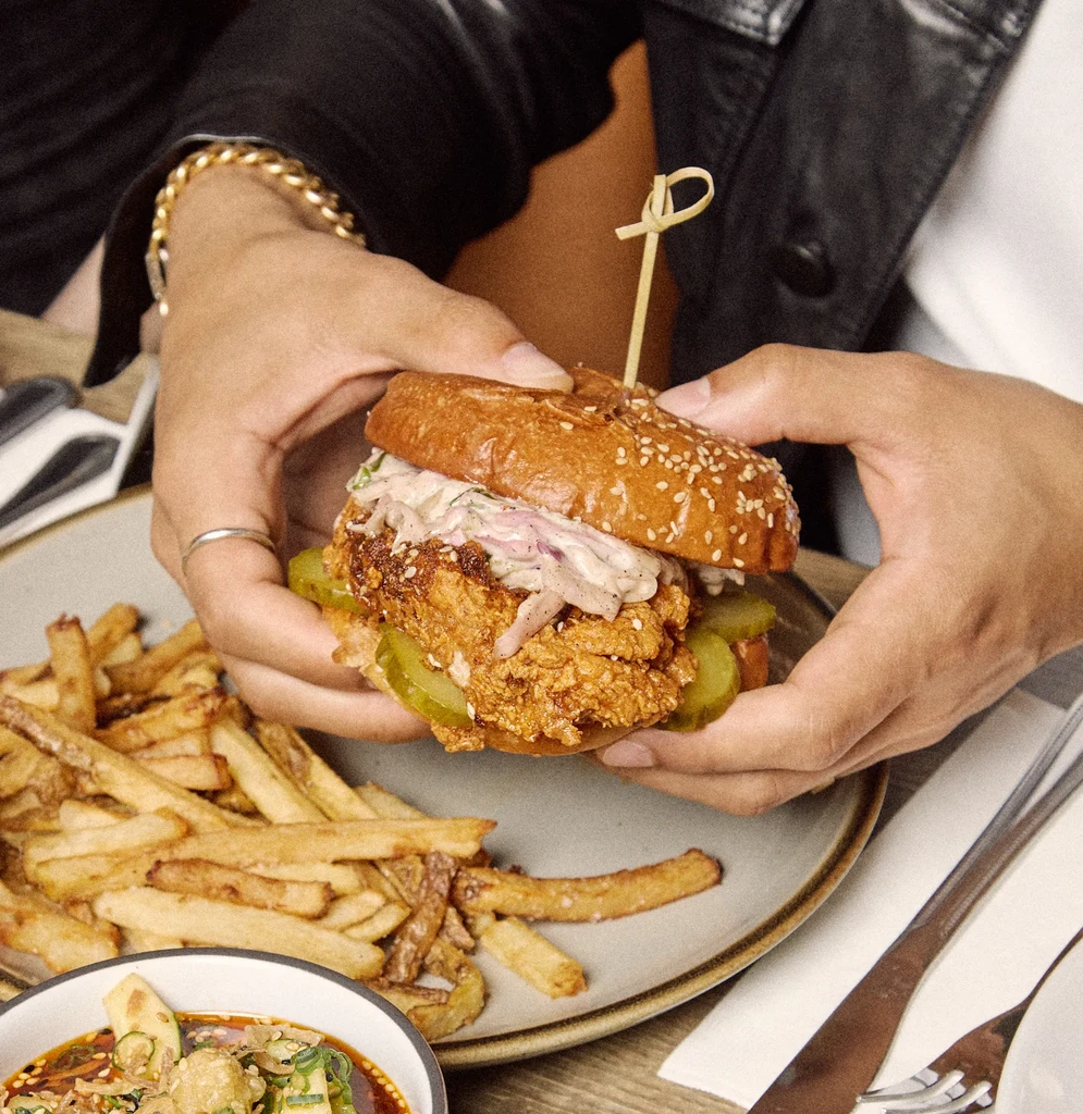 Hands hold a crispy fried chicken sandwich with fries and coleslaw.