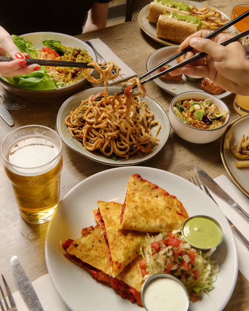 Overhead: Hands sharing noodles, quesadillas, salad, and beer at a table.