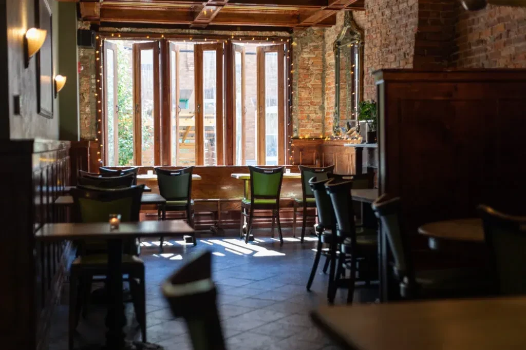 Empty restaurant dining room with wooden tables and large sunlit windows.