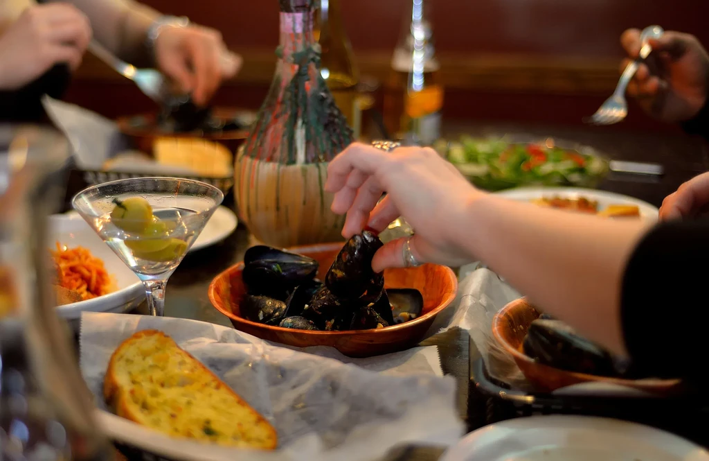 Hands reaching for mussels, martini, and garlic bread on a dining table.