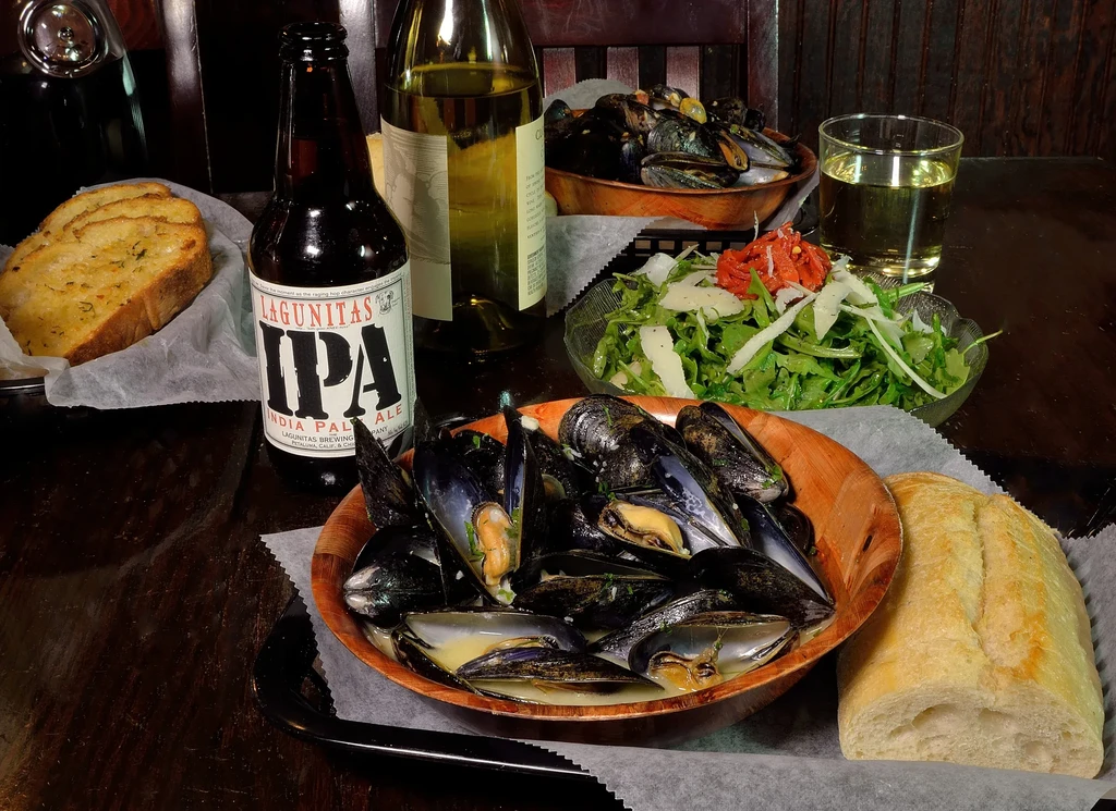 Mussels, garlic bread, beer, wine, and salad on a rustic wooden table.