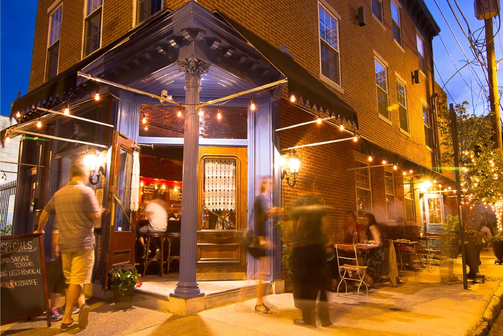 Evening view: brick restaurant, string lights, blurred people, chalkboard sign.