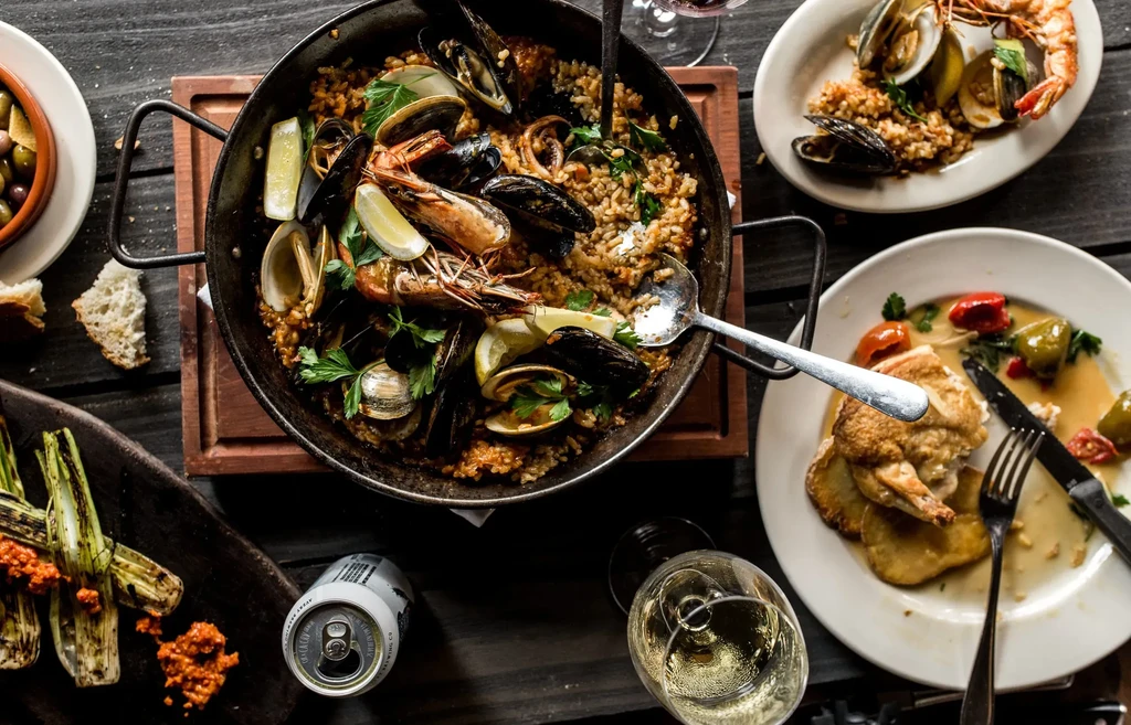 Overhead view: Rustic seafood paella, side dishes, and drinks on a table.