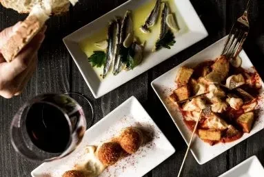 Table laden with tapas: marinated fish, potatoes, croquettes, wine, bread.