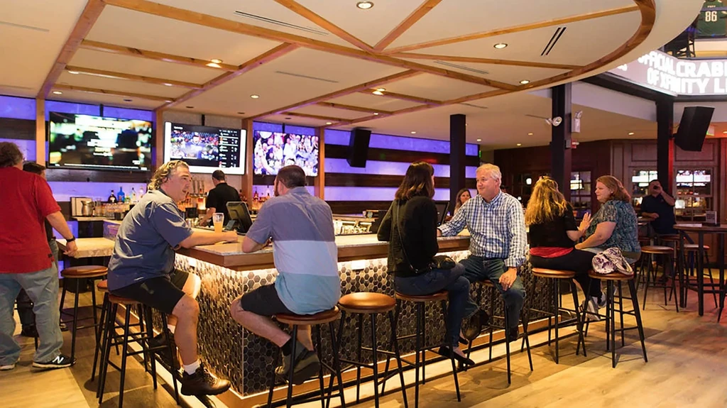 Lively sports bar interior with patrons, hexagonal bar, and sports on TVs.