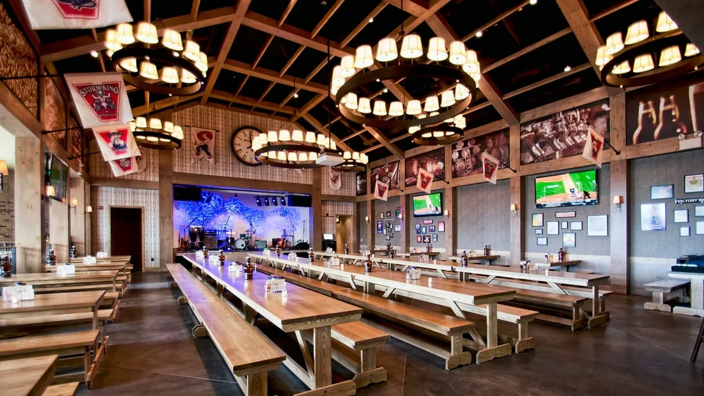 Empty rustic beer hall with high ceilings, long tables, and a stage.