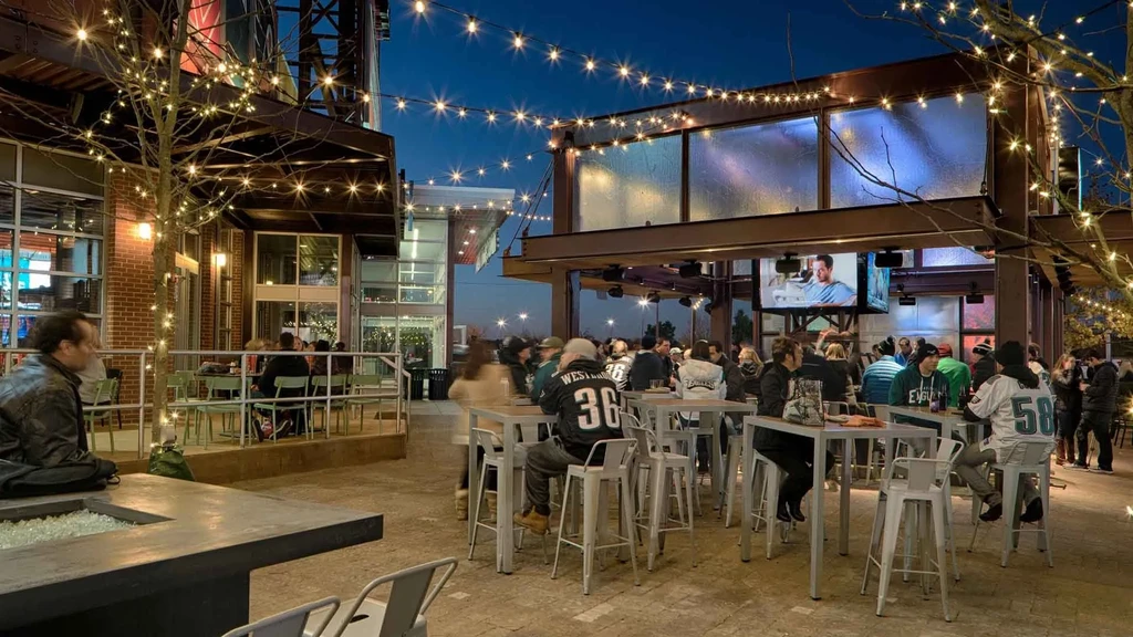 Outdoor patio at night with string lights, people, and TVs.