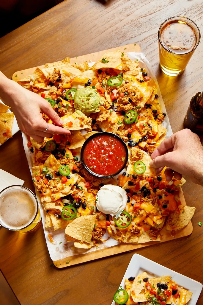 Two hands sharing a platter of loaded nachos with dips and beer.