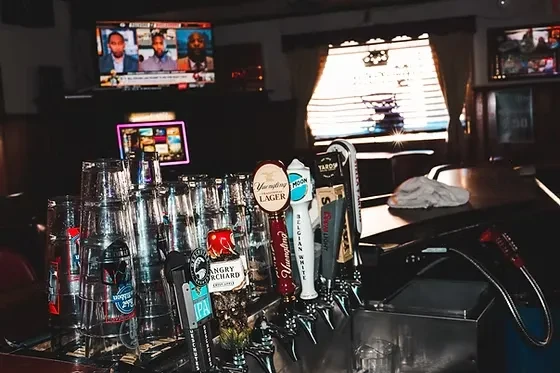 Bar counter with multiple beer taps, empty glasses, and various screens.