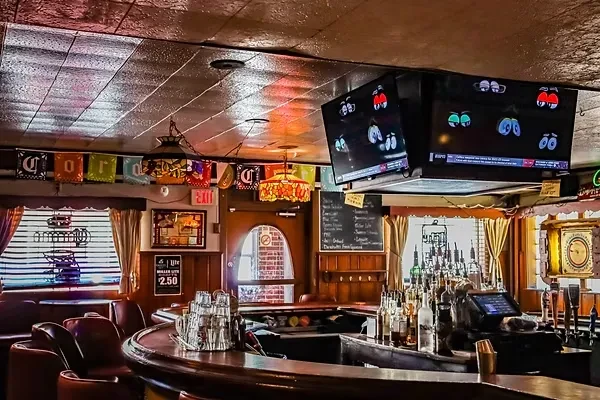Dimly lit bar interior with a wooden counter, liquor bottles, TVs, and seating.