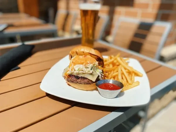 Burger with creamy topping, golden fries, ketchup, and beer outdoors.