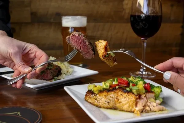 Two hands fork steak and salmon dishes with beer and wine.