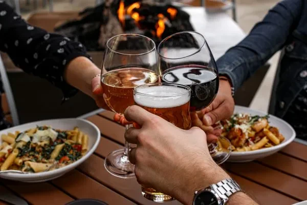 Three hands toast with wine and beer by an outdoor fire pit.